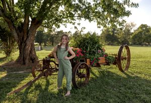 Casual outdoor portrait with antique farm equipment and lush garden backdrop.