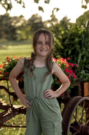 Child in olive green dress poses playfully in garden setting with red flowers and wooden decor.