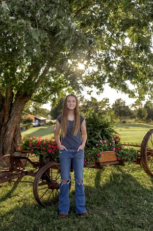 Young person in navy top and jeans stands near blooming red flowers and rustic wagon wheel.