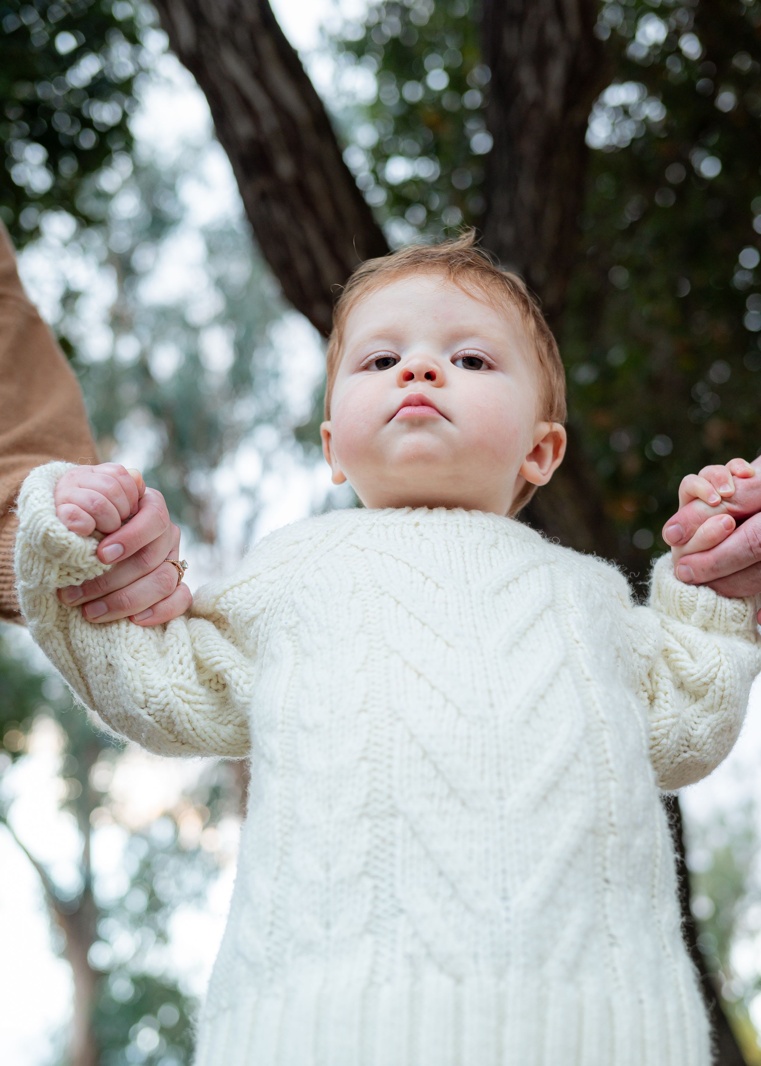 Baby in white knit sweater looking up while being held outdoors.