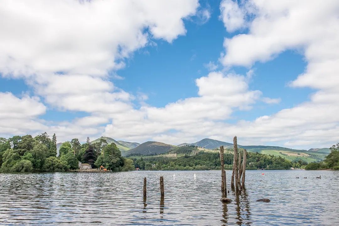 Calm lake with old wooden posts and distant mountains under a bright blue sky.