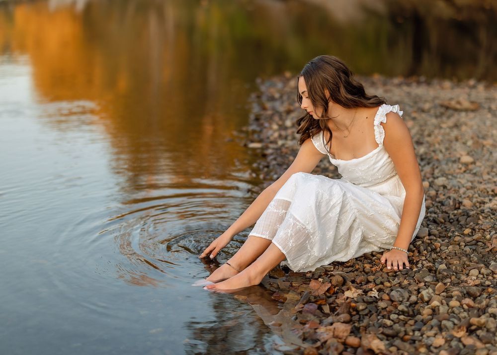 White dress flowing as someone sits along a rocky riverbank during a golden hour photoshoot.