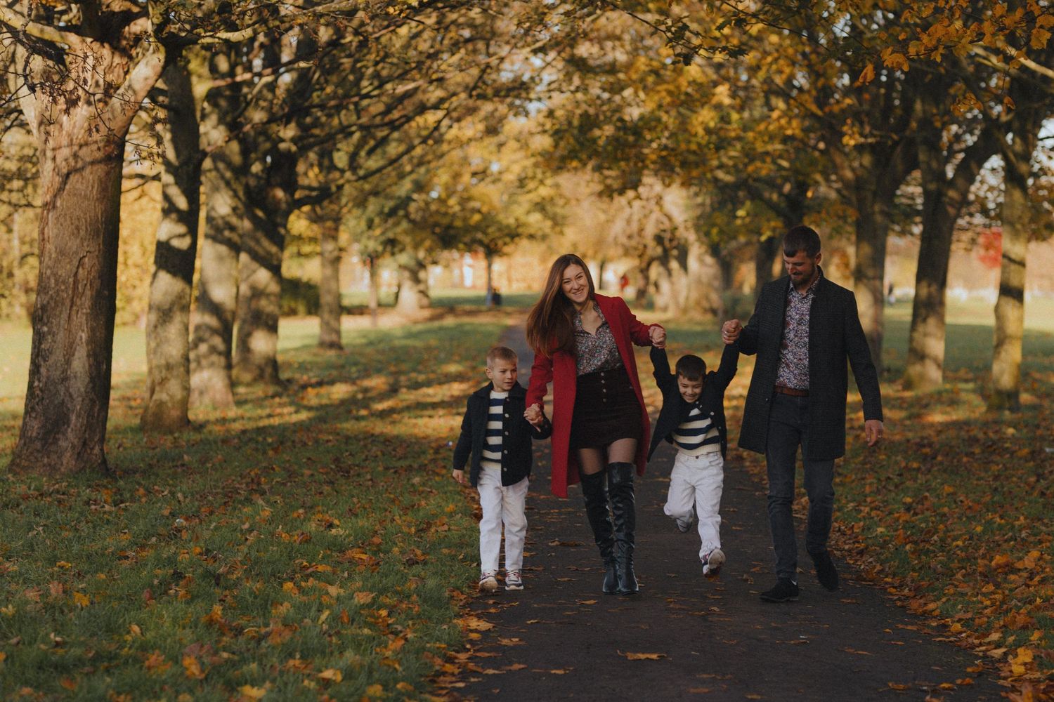People walking along a tree-lined path covered in autumn leaves during golden hour.