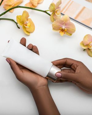 Hands holding a white cosmetic tube with metallic cap, surrounded by orchids and magazine skincare product photography in Spain, Europe, EU