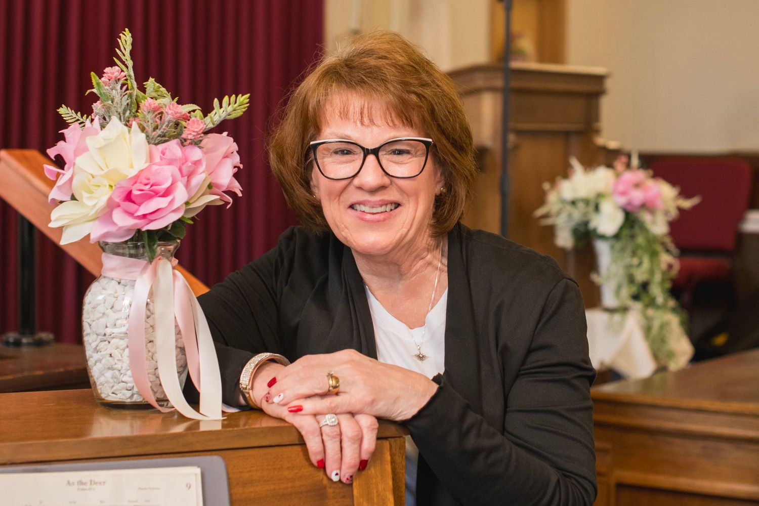 Smiling woman with glasses leaning on wooden surface with floral arrangement nearby.