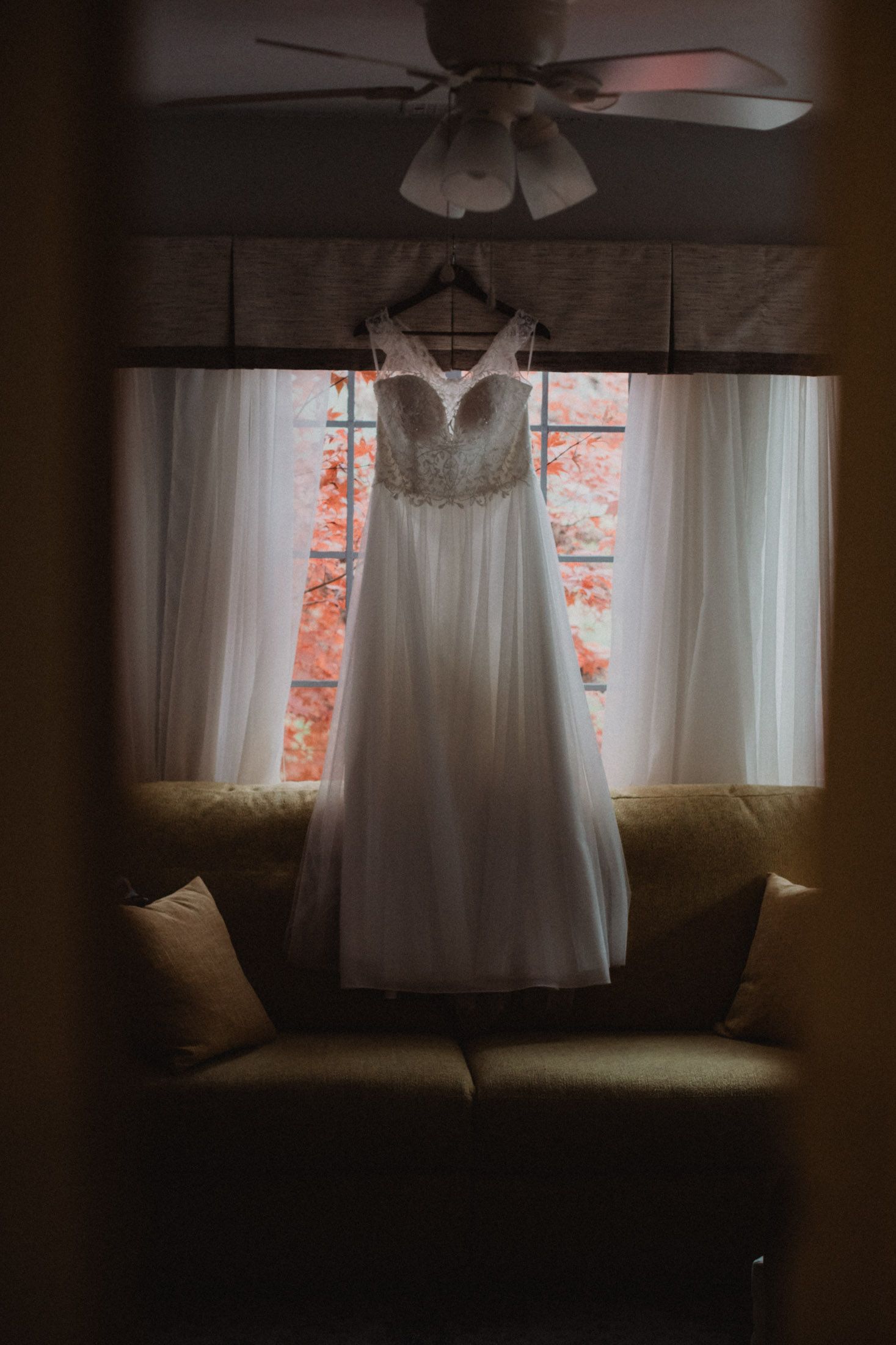 White wedding dress hanging in front of window with soft natural light and autumn foliage visible outside.