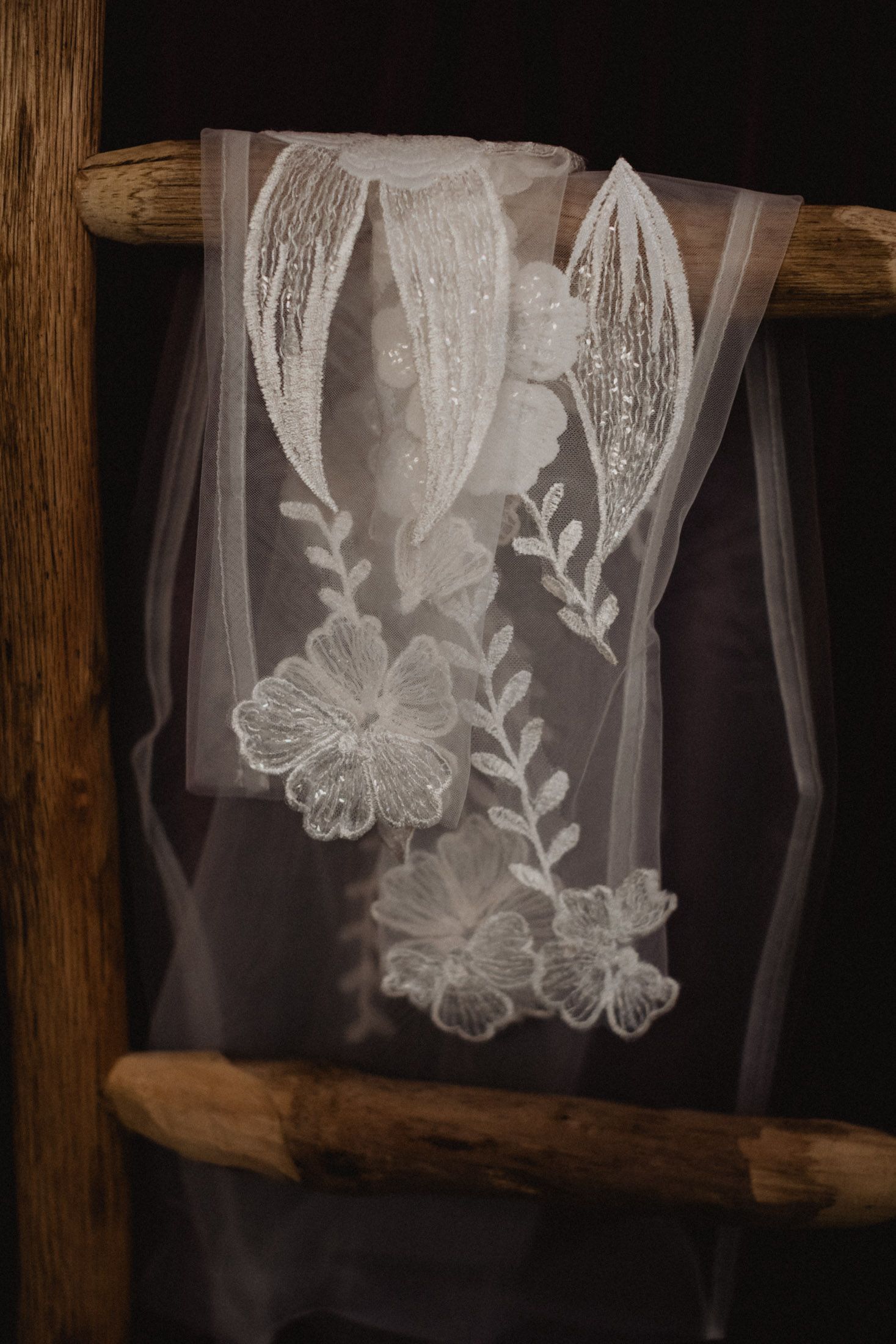 Delicate white lace veil with floral embroidery hangs on rustic wooden ladder against dark background.