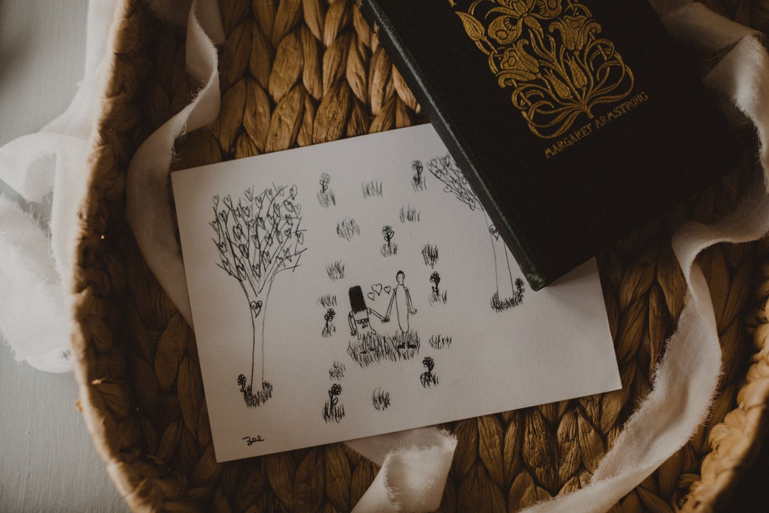 Simple black and white sketch of figures and trees on white paper next to a black book on woven surface.