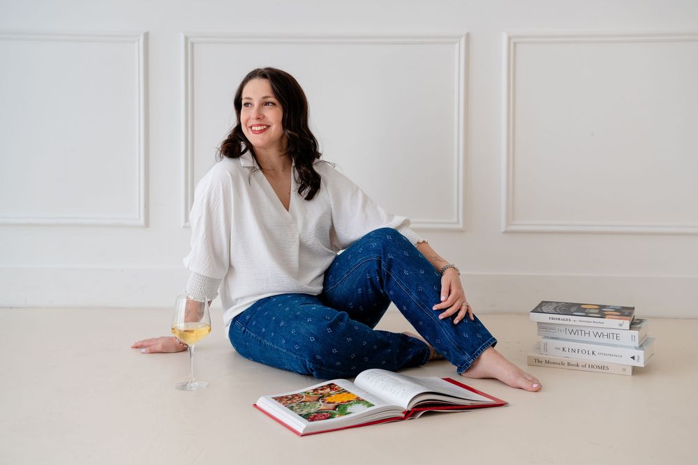 Person in white sweater and jeans lounges on floor reading colorful book against white wall.
