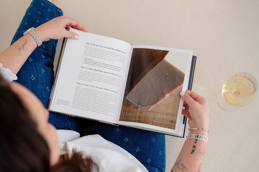 Someone reading a hardcover book while holding a glass of white wine on a blue couch.