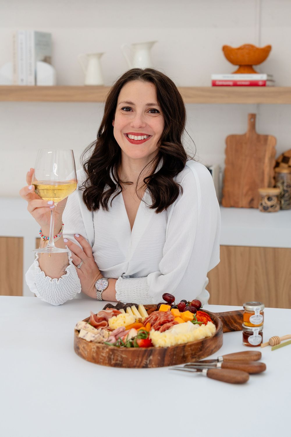 A charcuterie board with cheese, fruits and meats is displayed on a white counter with wine glasses.