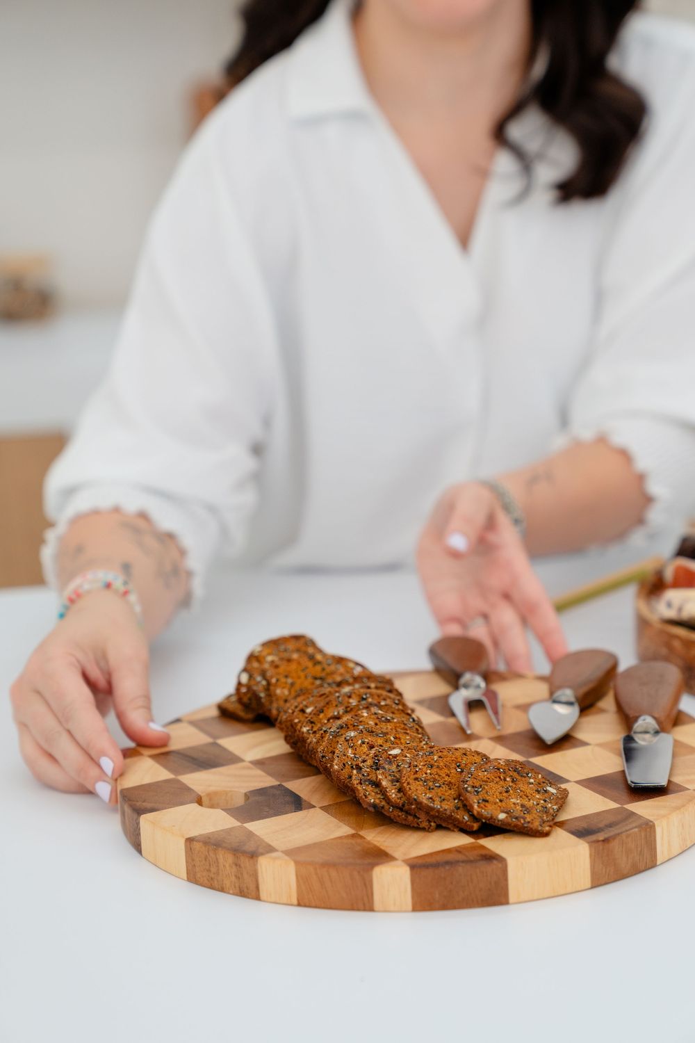 Hands arrange food items on a decorative wooden checkerboard cutting board in a bright kitchen setting.