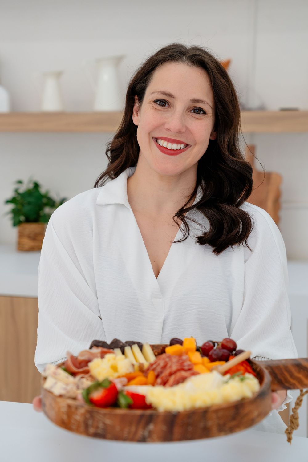 Woman in white bathrobe smiles while enjoying a colorful charcuterie board spread with meats, cheeses and fruits.