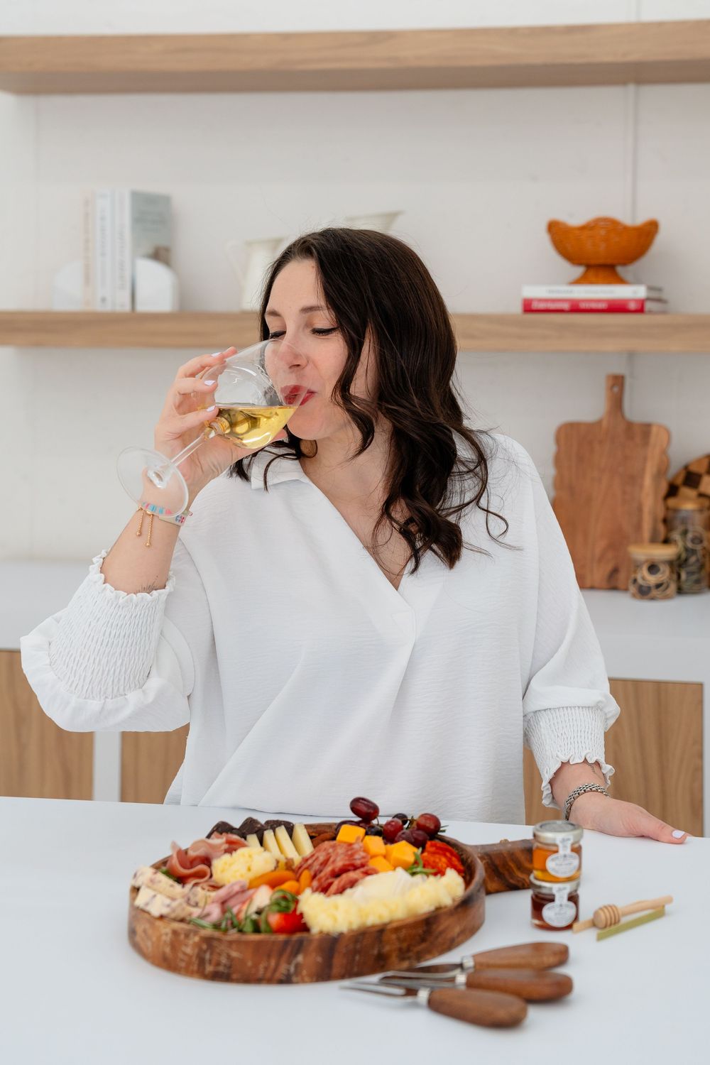 A charcuterie board with meats, cheeses and fruits is enjoyed in a bright modern kitchen with wooden shelving.