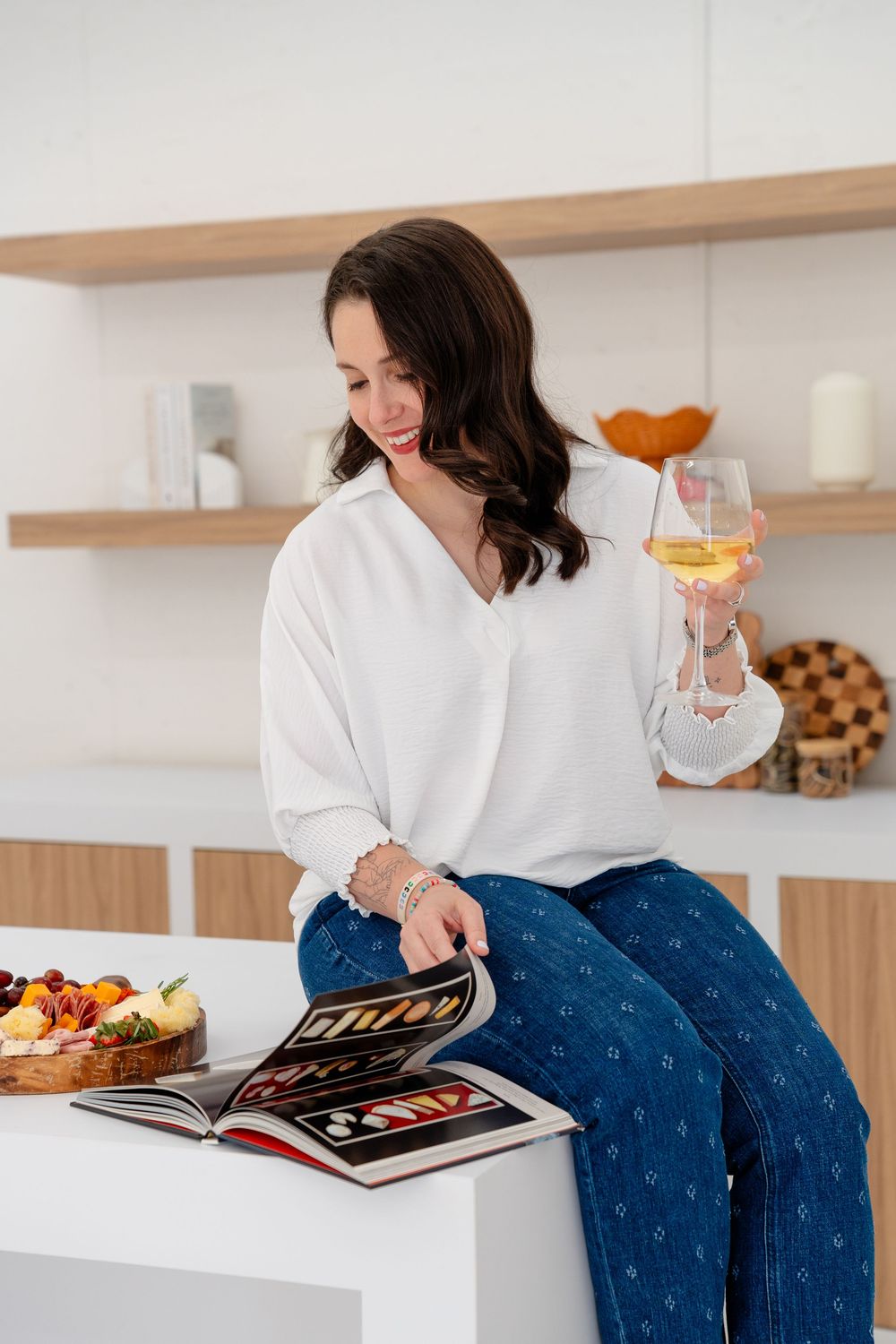 Someone in a white shirt and patterned jeans relaxes with a book and snacks on a modern white couch.