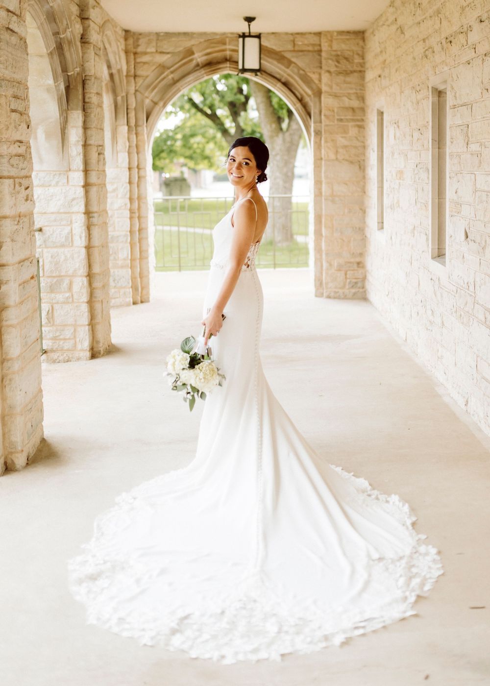 A bride in a white wedding gown poses elegantly in a stone archway corridor.