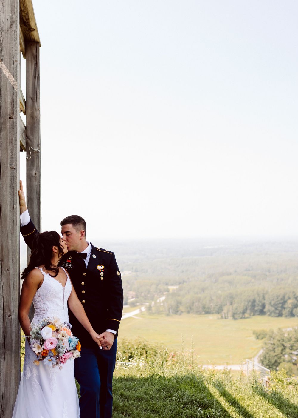 Wedding couple sharing intimate moment against wooden posts with scenic valley view.