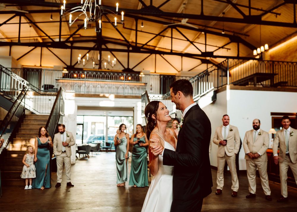 Newlyweds share their first dance in an industrial-style venue with exposed beams and wedding party looking on.