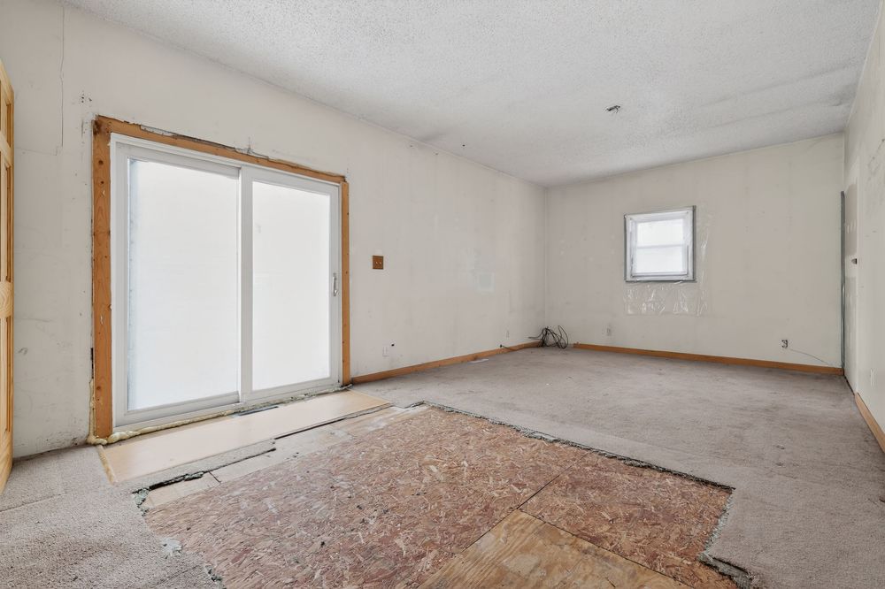 Empty room with sliding glass door, neutral walls, and subfloor exposed during renovation.