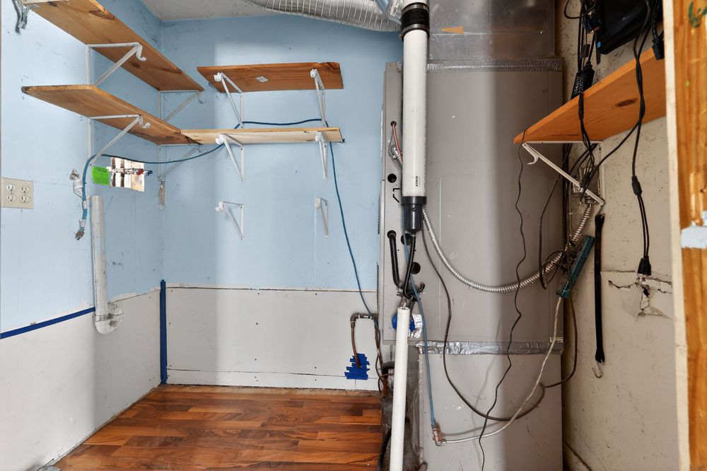 Utility area with tankless water heater, sump pump, exposed pipes, and wooden shelving.