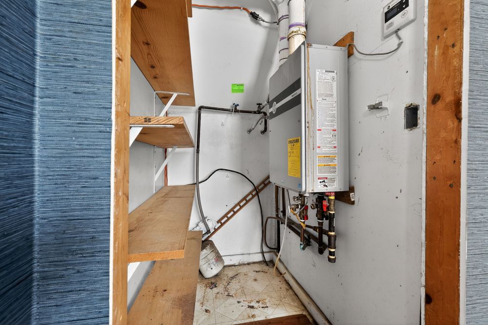 Narrow utility closet with tankless water heater, exposed pipes, and wooden shelving.