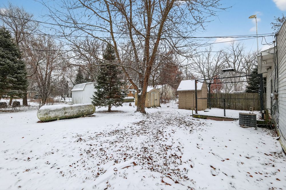 Snow-covered backyard with mature trees, wooden shed, and fence in winter landscape.