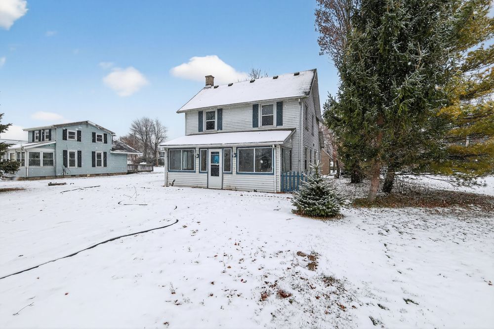 Snow-covered front yard of a two-story white farmhouse with a screened porch and evergreen tree in winter.