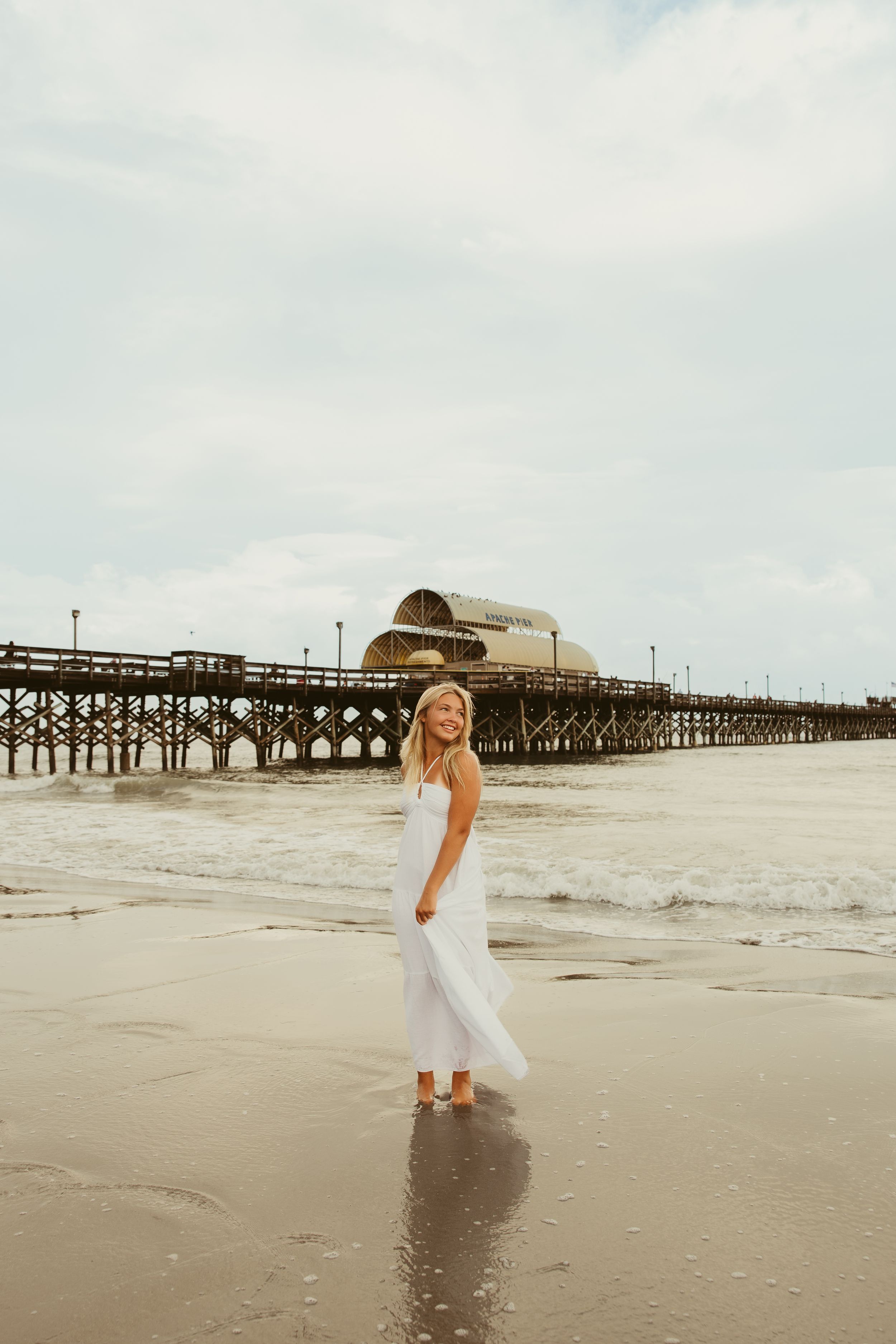 Smiling blonde girl in white dress walking on beach with wooden pier in background.