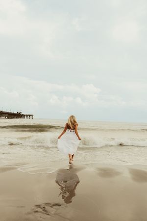 Woman in white dress walking toward ocean waves on a sandy beach with a pier in the background.