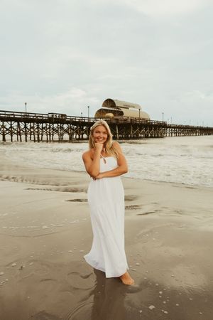Smiling blonde woman in white maxi dress standing barefoot on beach with wooden pier behind her.