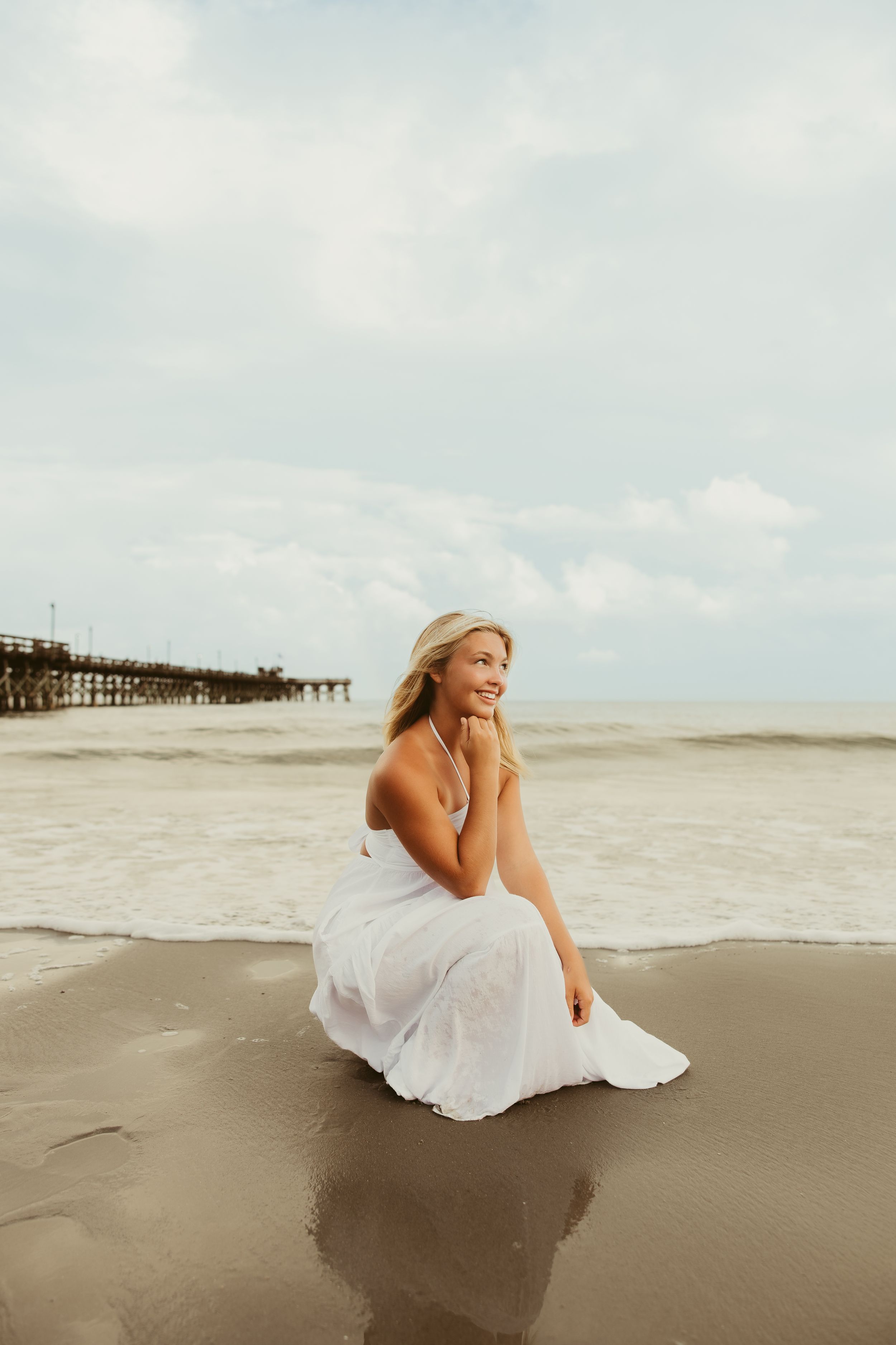 Blonde woman in white dress sitting on wet sand at beach smiling with ocean waves and pier nearby.