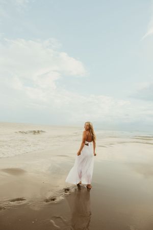 Woman in flowing white dress walking along reflective wet sand beach with cloudy sky overhead.