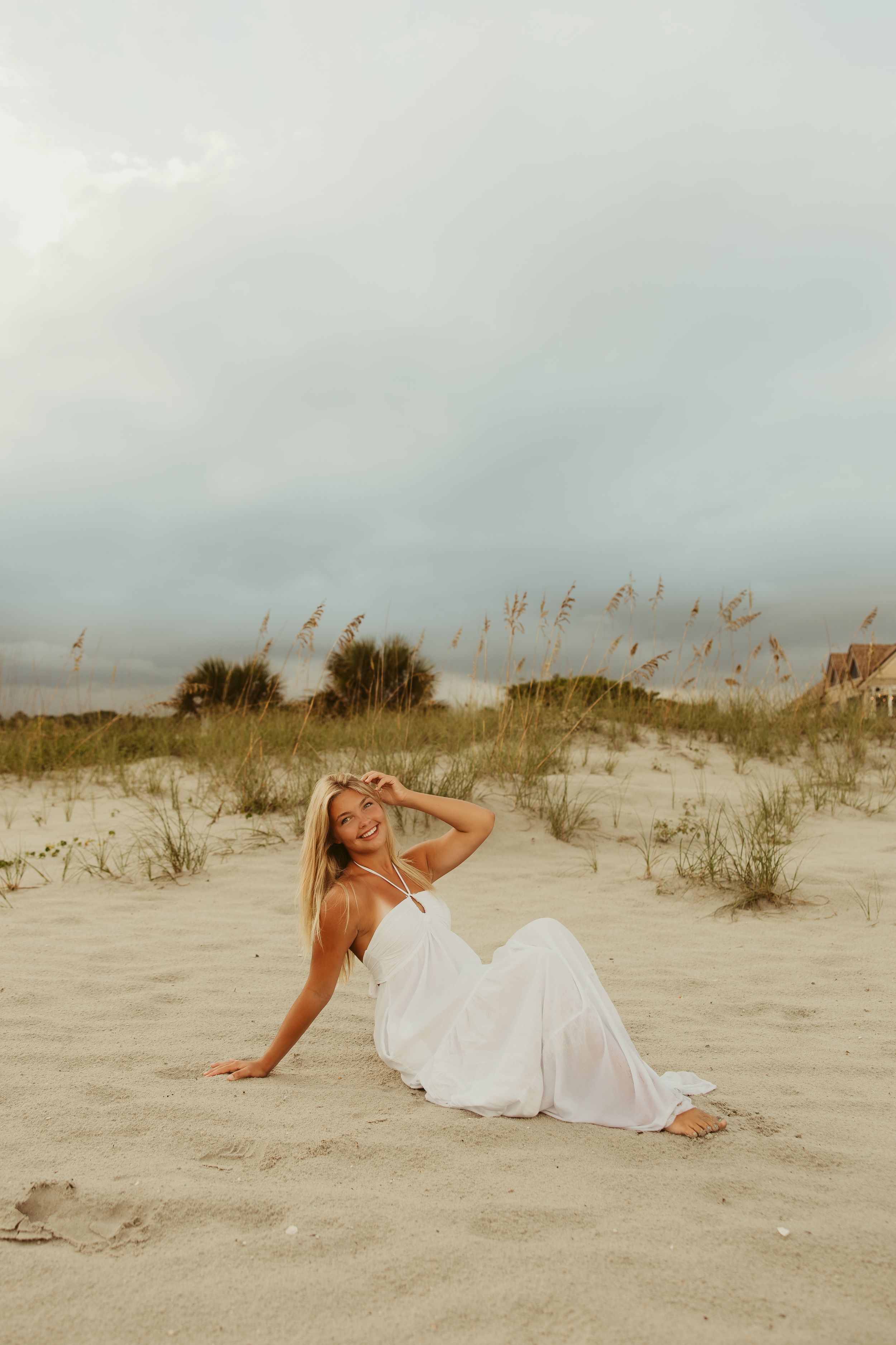 Blonde woman in white halter dress sitting on sandy beach dunes with sea oats and cloudy sky.