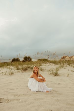 Smiling woman in white dress sitting cross-legged on sand dunes with beach grass and houses behind.