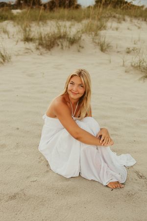 Young blonde woman in white dress sitting on sandy beach dunes smiling at camera on cloudy day.