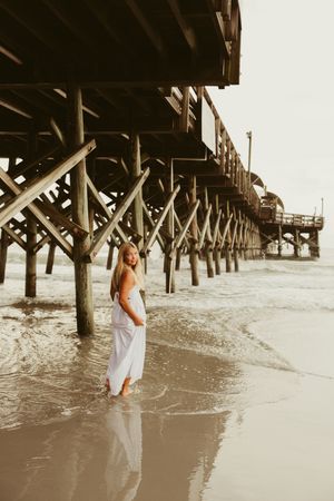 Blonde woman in white dress standing under wooden beach pier with ocean waves around her feet.