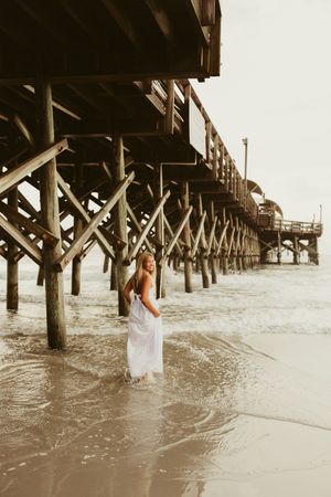 Woman in white dress standing beneath large wooden beach pier with waves washing around her feet.
