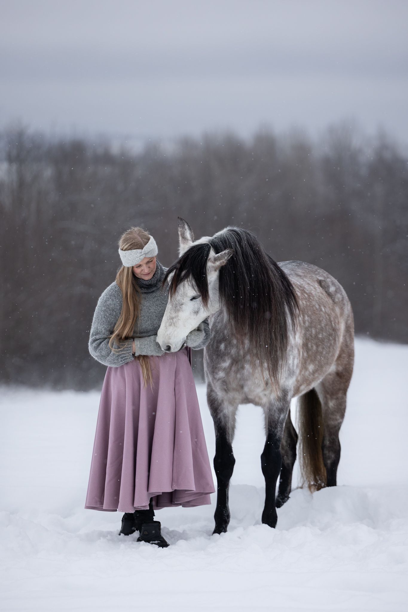 A person in a purple dress stands beside a dappled grey horse in a snowy winter landscape.