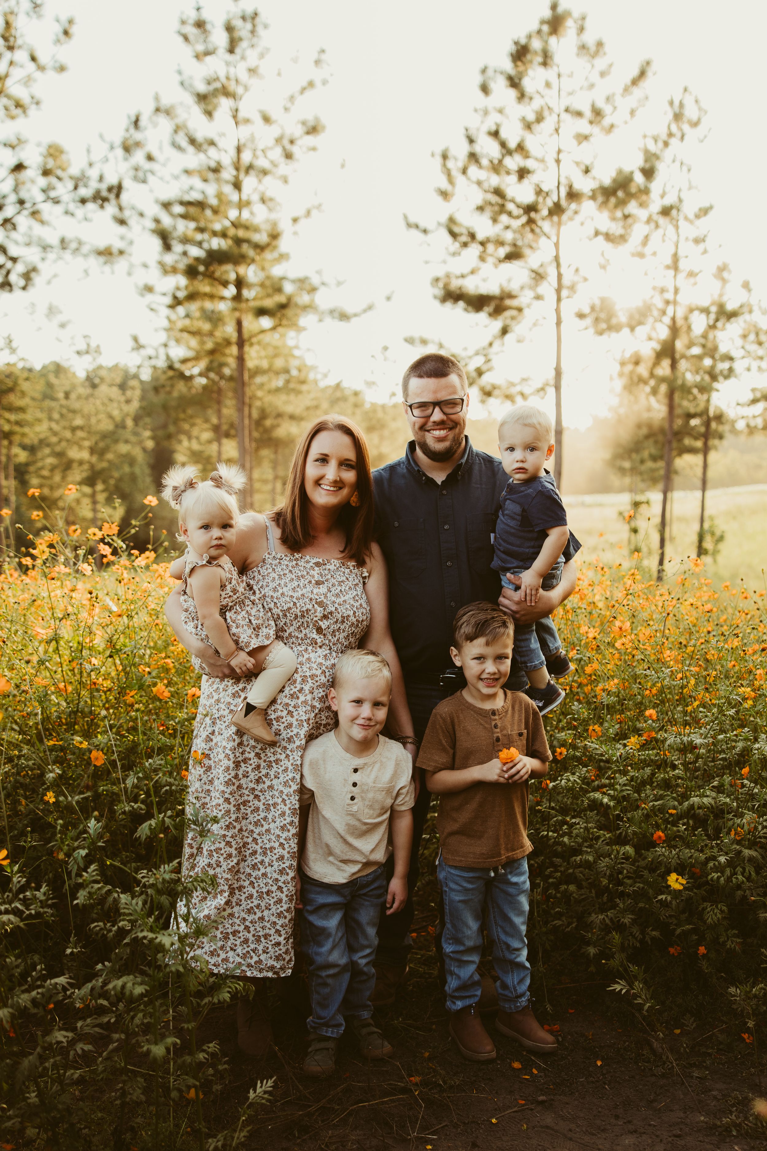 Happy family of six posing in a vibrant orange wildflower field surrounded by pine trees at sunset.