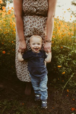 Smiling baby boy standing in a field of orange wildflowers, held by mother in floral dress.
