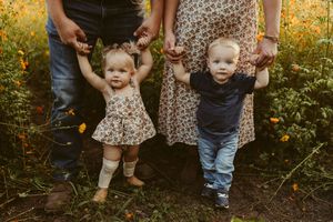 Two toddlers holding parents hands standing in a sunny wildflower meadow during golden hour.