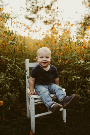 Adorable smiling baby boy sitting on white chair surrounded by orange wildflowers at sunset.