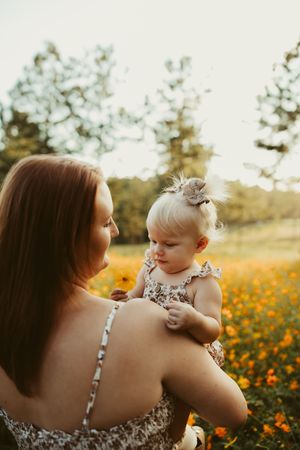 Mother holding her blonde toddler daughter in a sunlit orange wildflower field at golden hour.