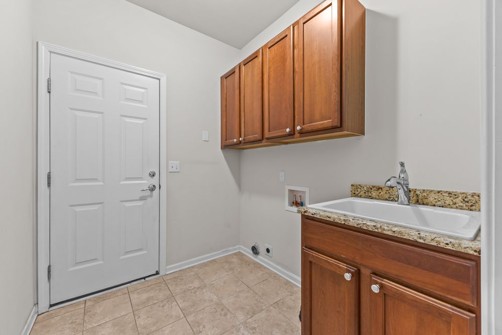 Laundry room with wooden cabinets, granite countertop, utility sink, and tile flooring near an exterior door.