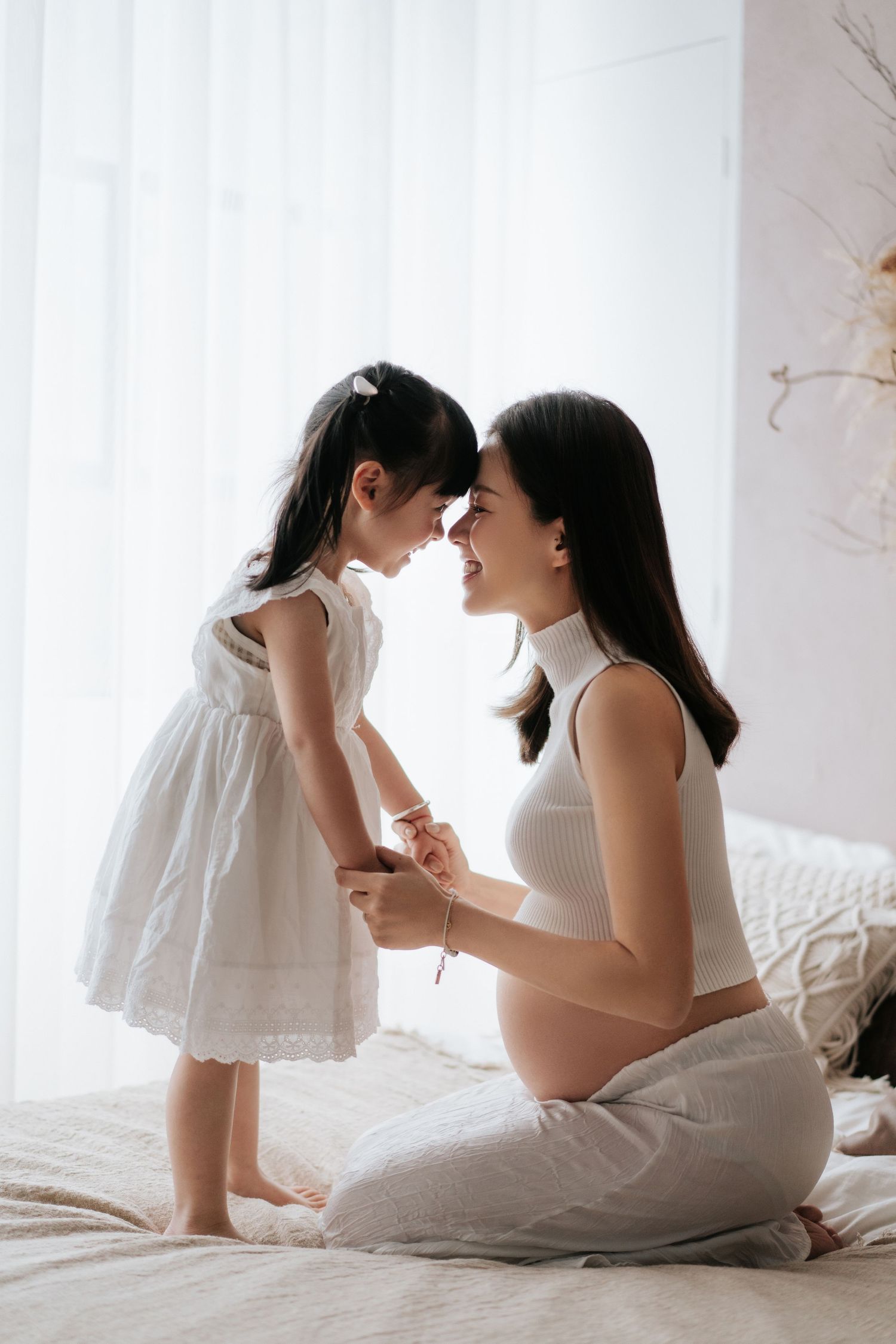 A tender moment between parent and child in white dresses sitting on a bed in soft natural light.