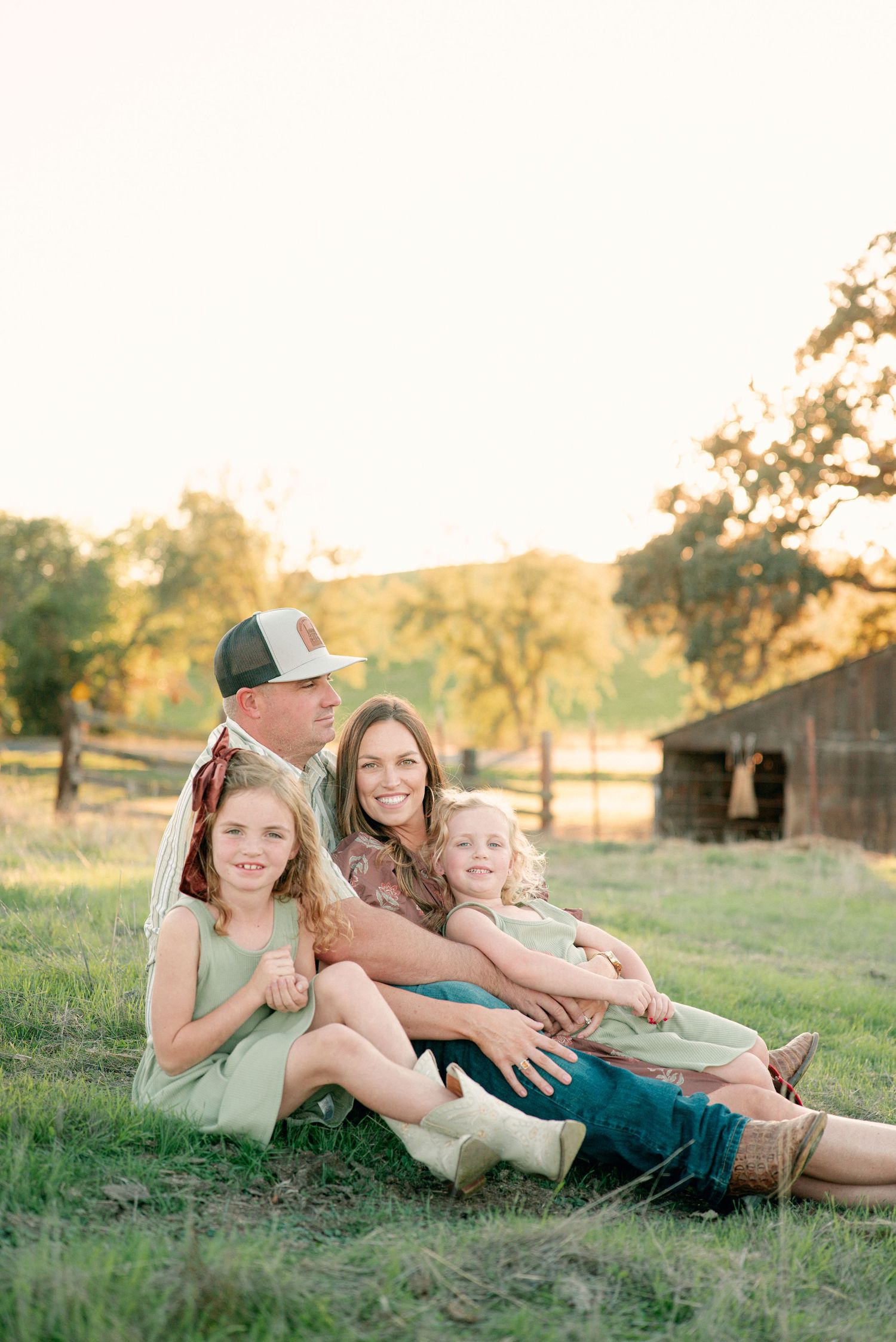 A family sits together on grassy field at sunset with rustic barn in background during golden hour photo session.
