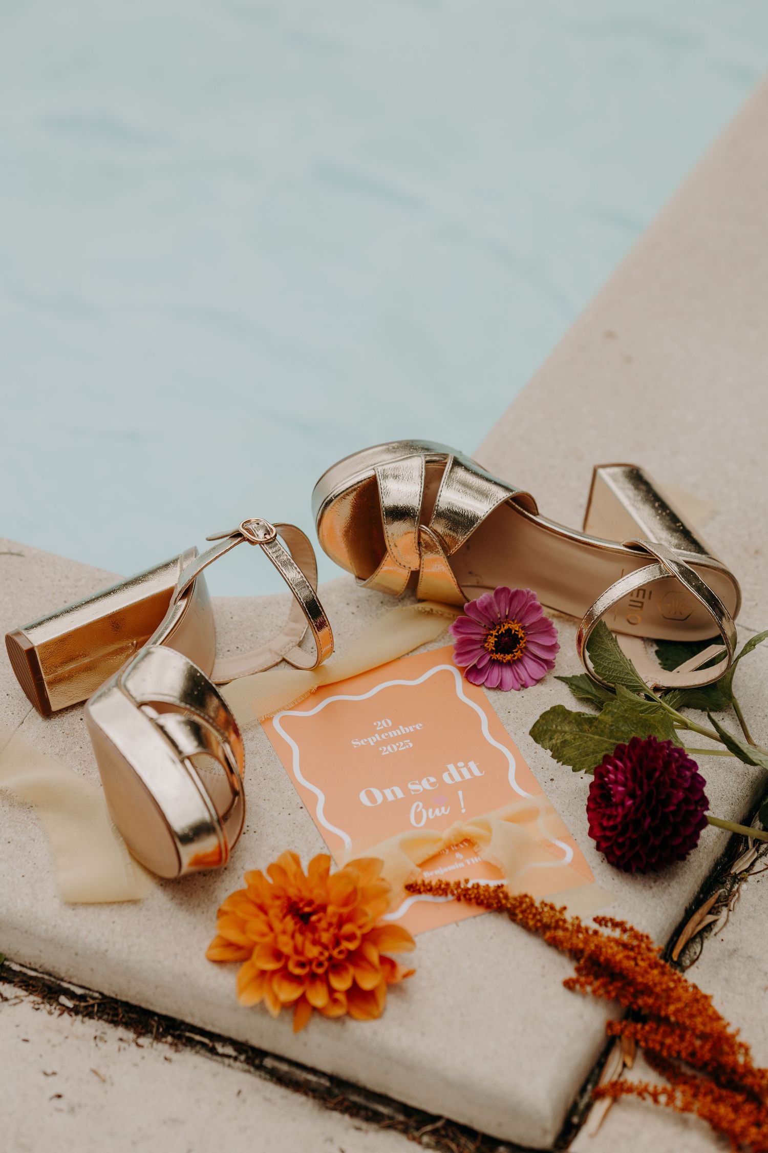Gold wedding shoes displayed with vibrant orange and purple dahlias on a neutral background.
