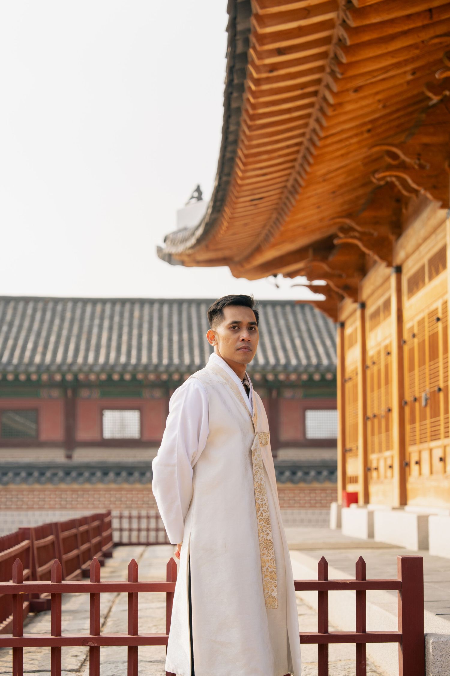 A person in traditional white Korean hanbok poses at a historic palace with traditional architecture in the background.