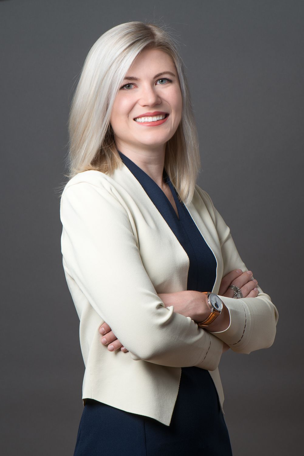 Professional in a white blazer and dark top stands with arms crossed against a gray background, smiling warmly.