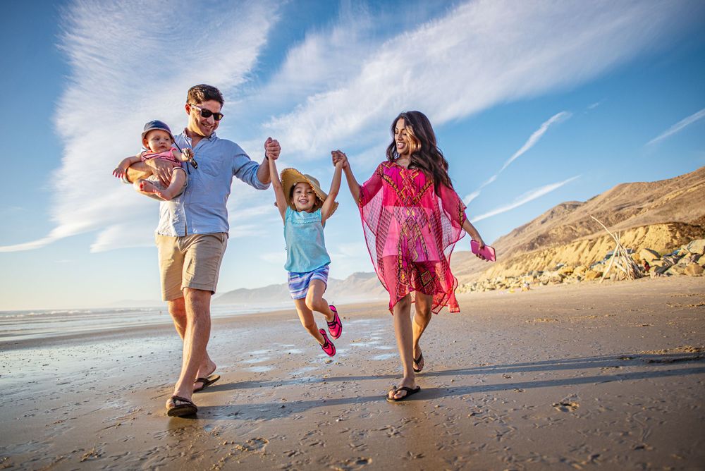 Family holding hands and jumping together while walking along a scenic beach at sunset.